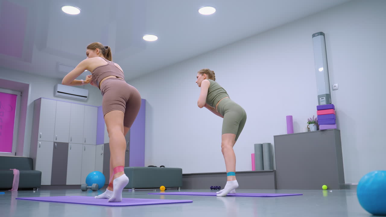 vista trasera de dos mujeres trabajando en esteras púrpuras usando bandas de resistencia en un gimnasio bien iluminado, realizan ejercicios de la parte inferior del cuerpo con la postura adecuada, rodeadas de equipos de fitness