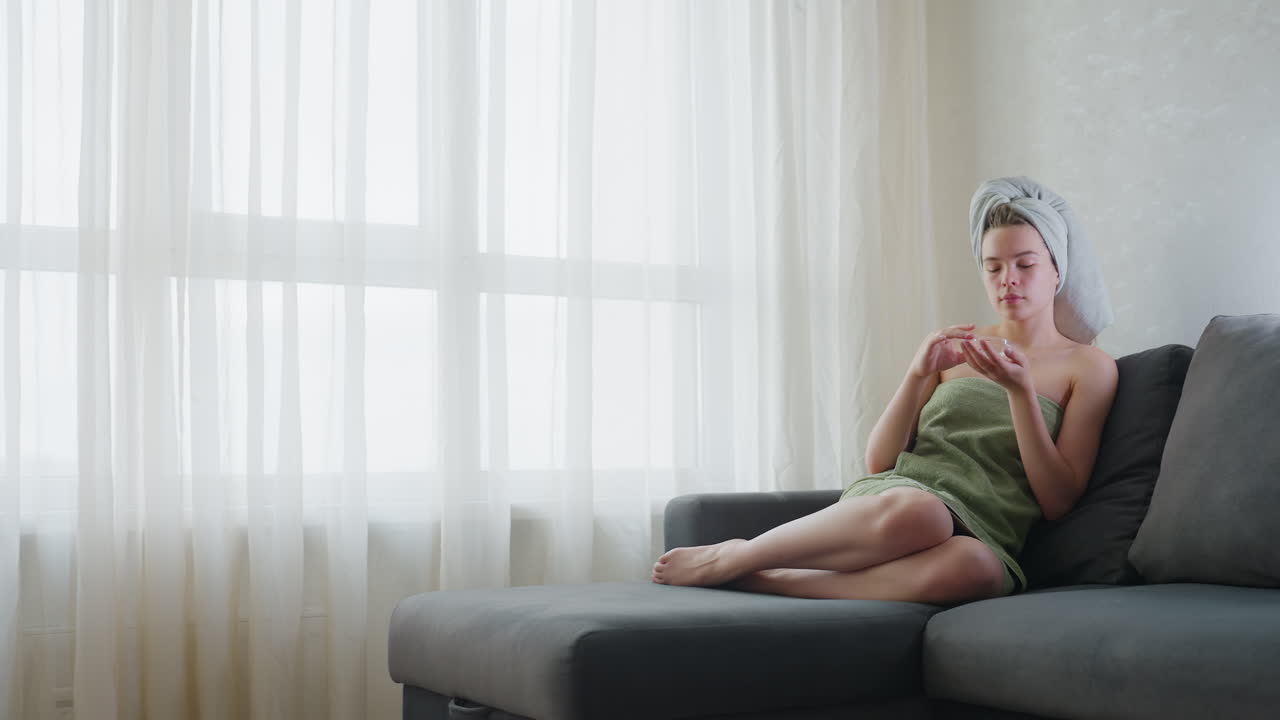 Lady wrapped in light green towel sitting cross-legged on couch beside large window covered with soft white cotton curtains gently applies liquid skincare substance to face in calm indoor setting