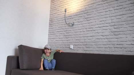 smiling caucasian toddler sitting in the corner of the sofa. lovely boy in a cap playing at home.