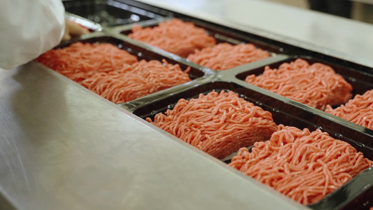 Food handler in a clean coat arranges fresh ground meat portions in black tray compartments on a metal work surface.