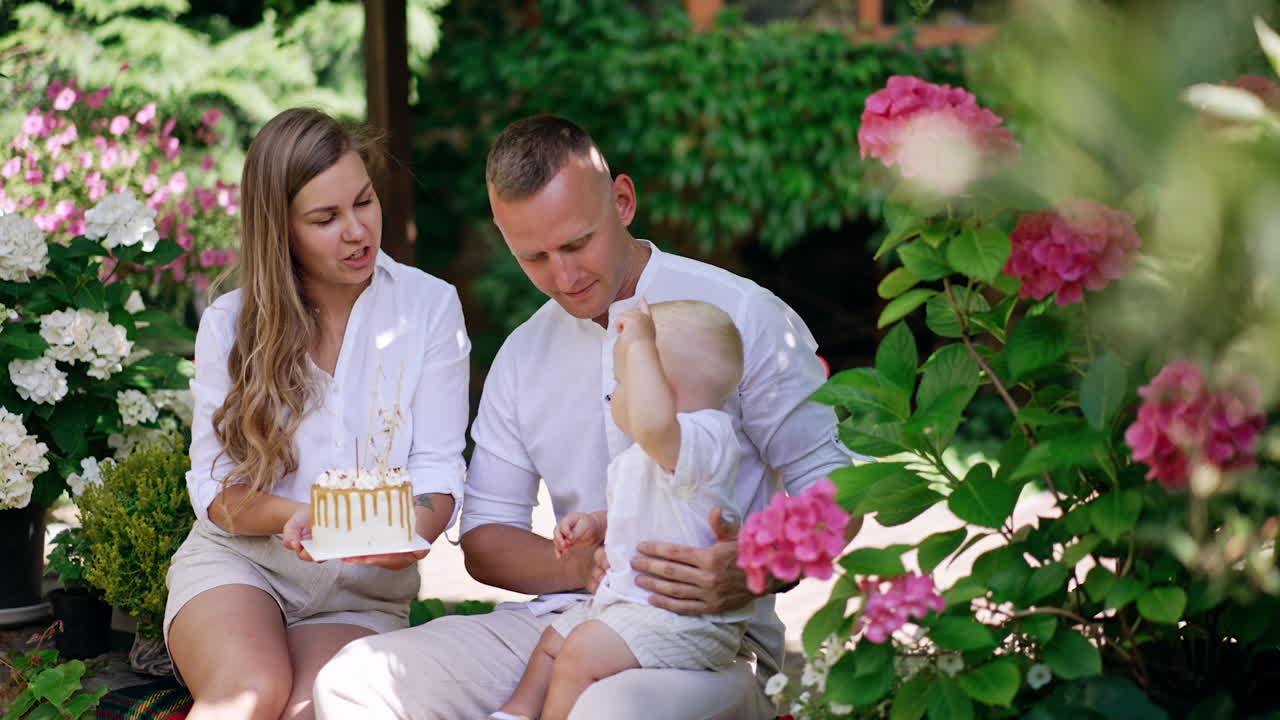 Happy mother and father with their baby outdoors. Mom is holding a cake and child tries to get it. Beautiful flowers around.