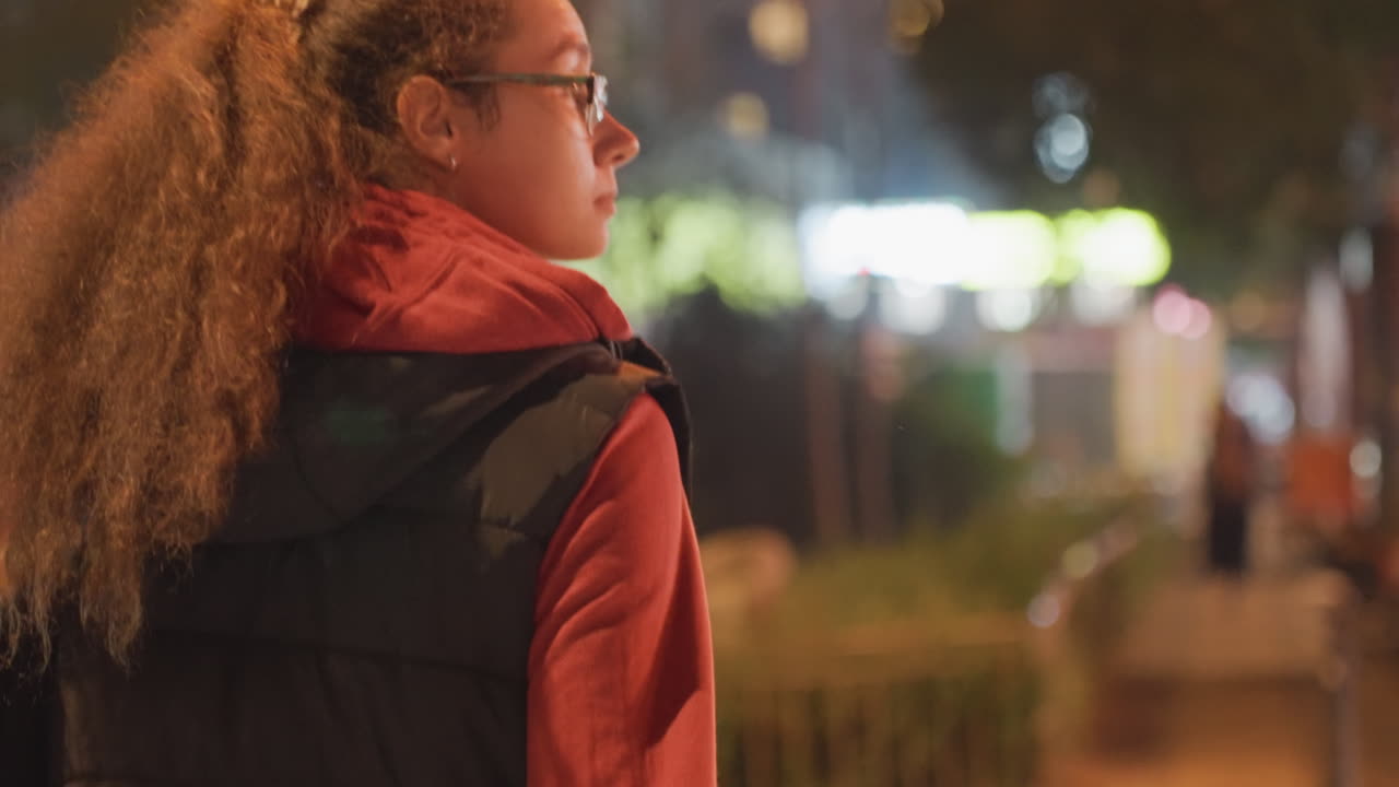 Young woman at crosswalk night backpack glasses long curly hair pulled into ponytail waiting under warm streetlights blurred traffic and neon signs create cinematic bokeh reflective commuter mood