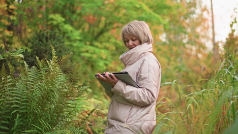 female researcher in beige jacket writing observations on tablet while conducting fieldwork in vibrant autumn forest, surrounded by tall grass and ferns, focused on plant data recording