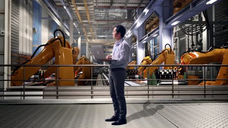 full body side view of an asian male professional worker with industrial robotic arms working, observes by looking up before he come to concentrating on the laptop and keep on typing