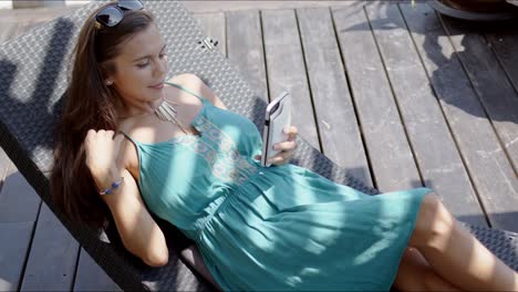 Woman-in-blue-dress-resting-in-lounge-chair-and-using-smartphone