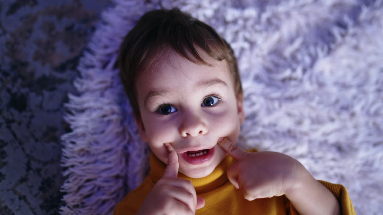 Charming Caucasian baby boy lies on the fluffy plaid. Cute toddler touches his cheeks, nose and ears smiling. Close up.