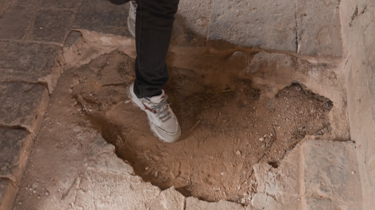 Crumbling stone floor with sinking hole person stepping near cavity closeup of sneaker entering loose sand and debris dramatic light beam and shadow tense movement and cautious balance archaeological