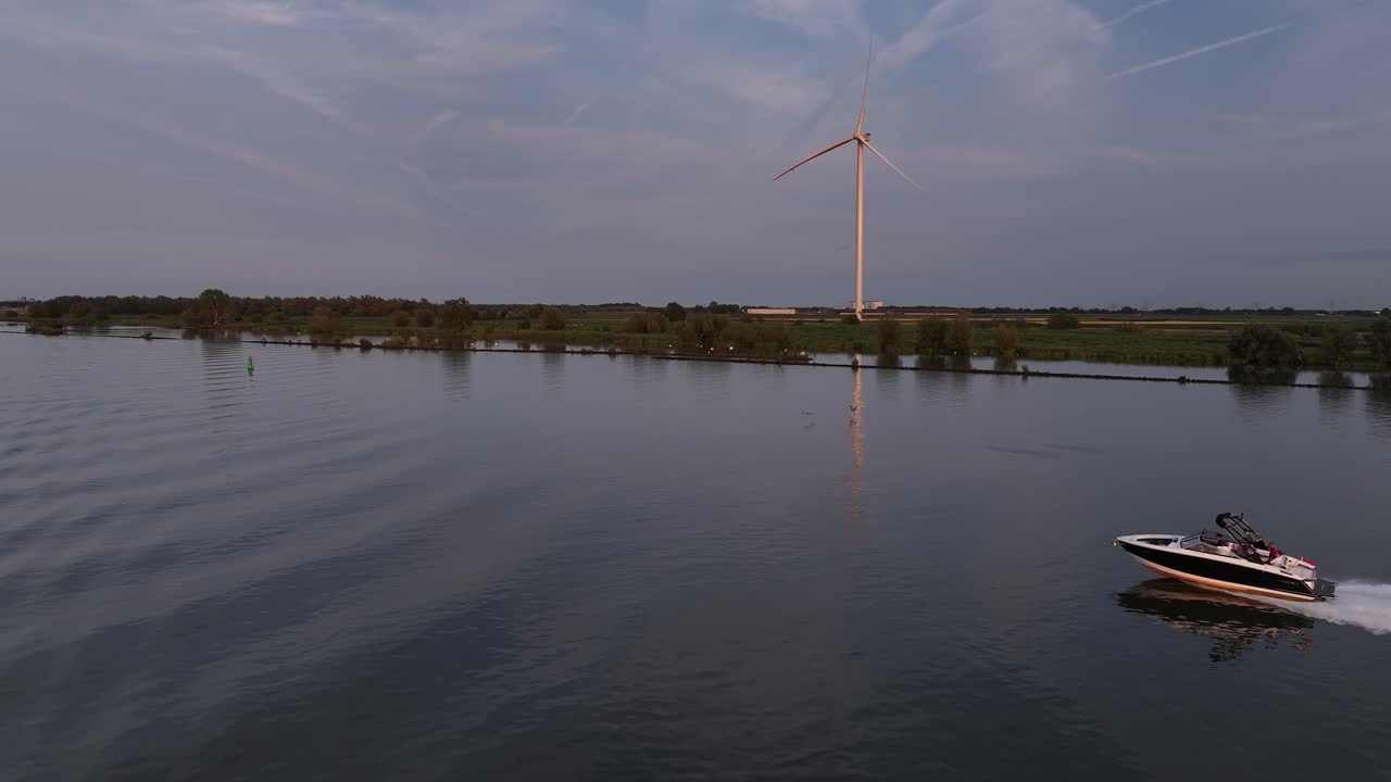 Motorboat travels fast on Dutch river by wind turbine, aerial follow pan