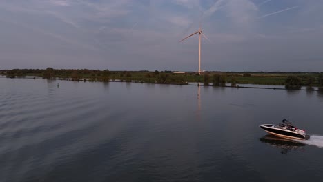 motorboat travels fast on dutch river by wind turbine, aerial follow pan