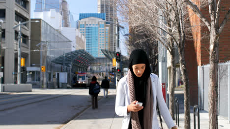 Vista-Frontal-De-Una-Joven-Asiática-Con-Hijab-Usando-Un-Teléfono-Móvil-En-La-Ciudad-4k