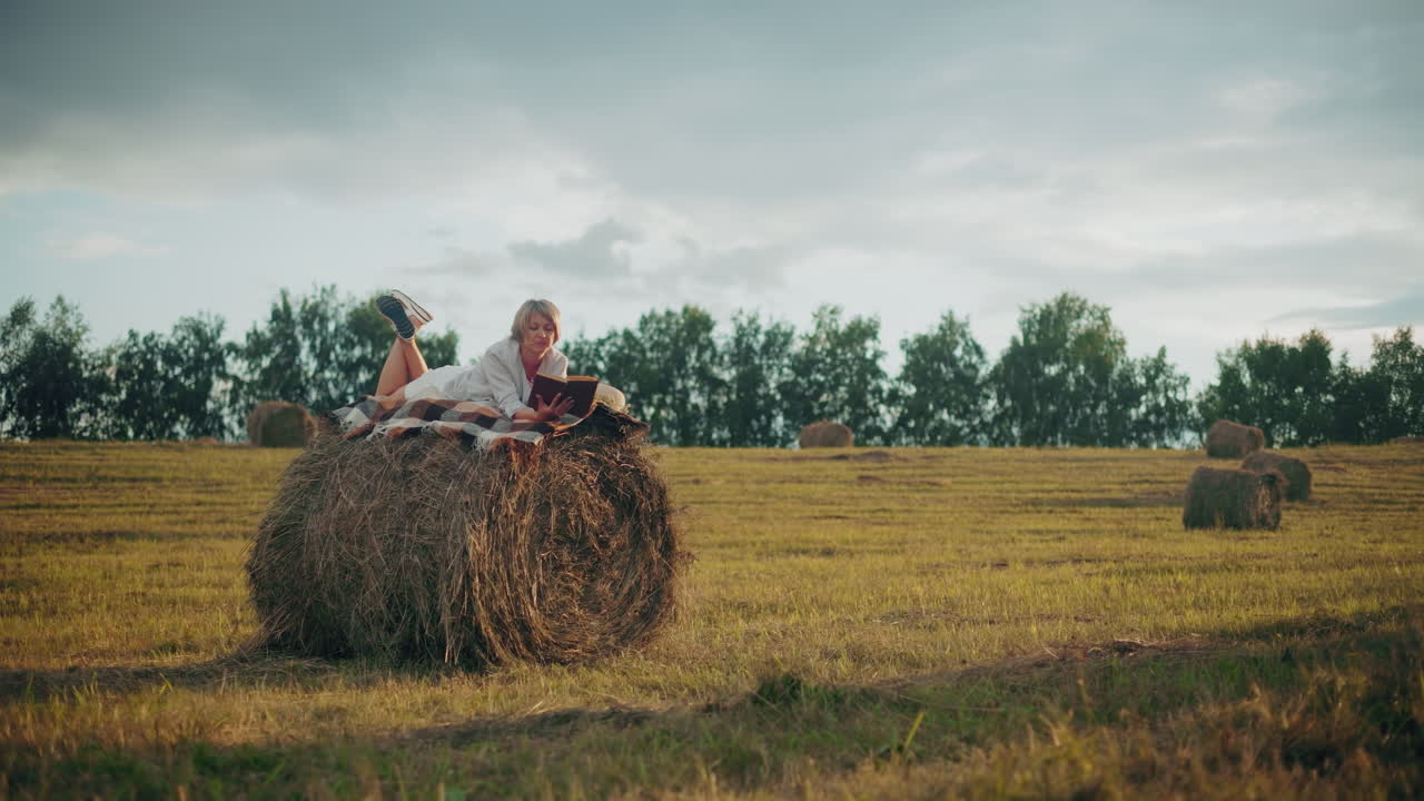 Dame slapper av med benet hevet på en høyballe med et rutete teppe, leser bok på et åpent jorde, nyter naturens ro med den fredelige utsikten over jordet rundt.