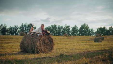 lady relaxing with leg raised on hay bale with checkered blanket, reading book in an open field, enjoying the serenity of nature with the peaceful surrounding view of the field