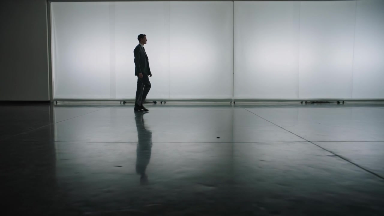 Entering left, suit-clad man walking toward center of hall, shiny floor, backlit wall, copy space