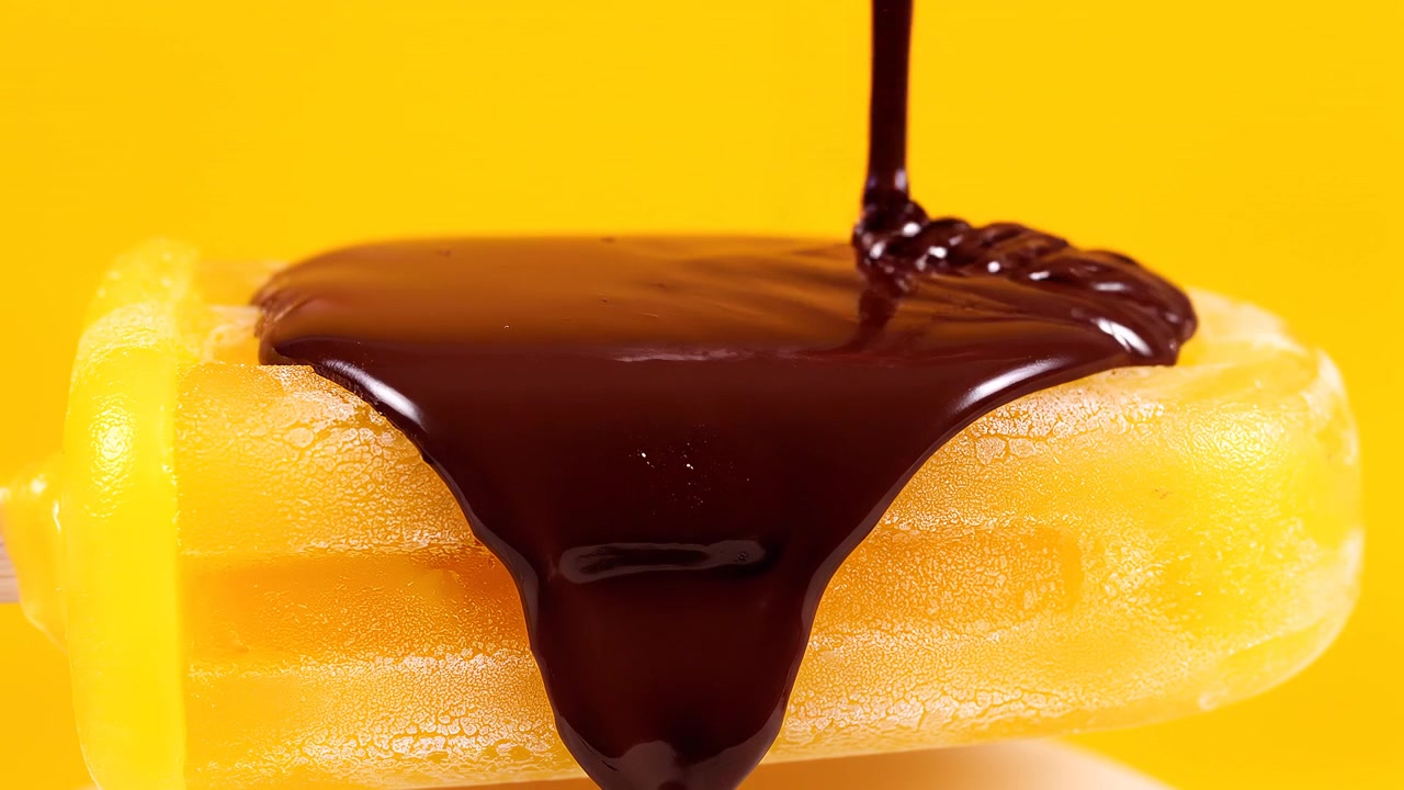 Salsa di cioccolato fondente fuso che cola su un ghiacciolo giallo.
