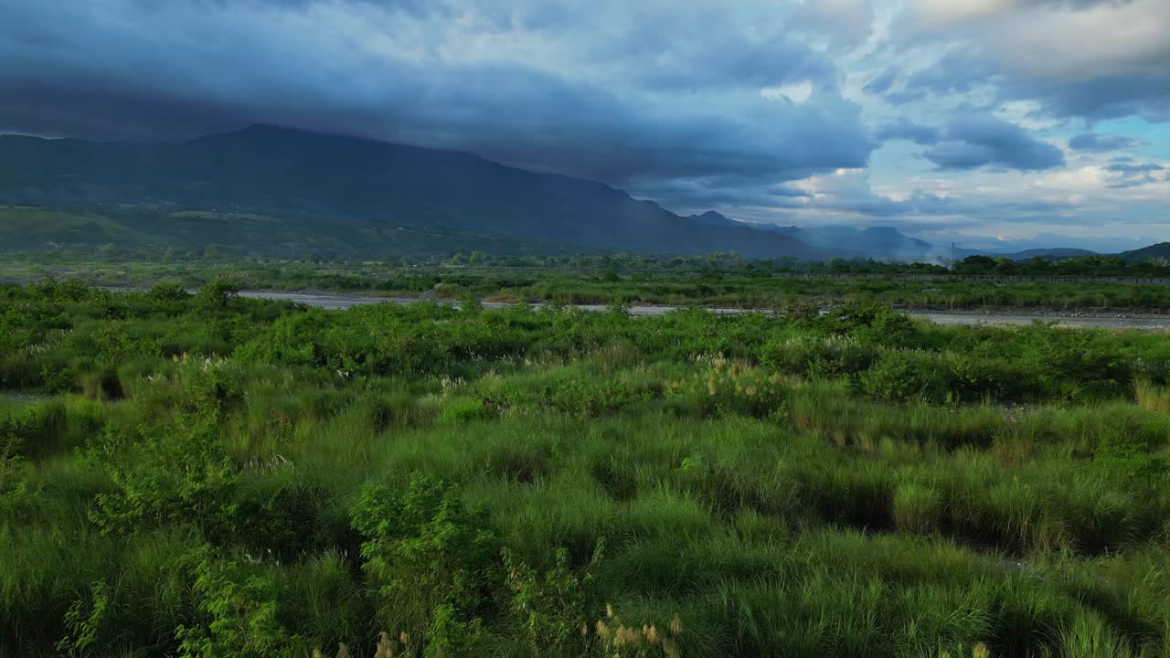 A forward ground aerial of grass fields near the Chico River reveals distant mountains shaded under clouds Bontoc, Nueva Vizcaya, Philippines