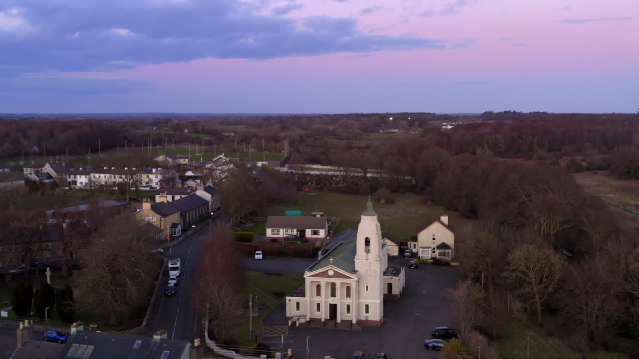 Town of Clarinbridge under a sky full of vibrant pink and purple sunset clouds from above, aerial establishing around church