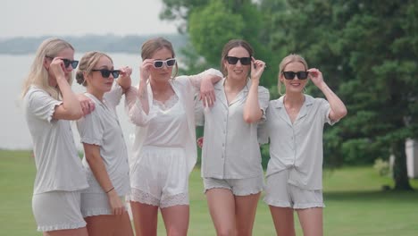 bridesmaids in white pajamas stand together outdoors by a lake, smiling and wearing sunglasses