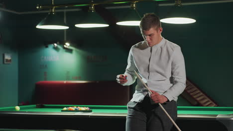 young man in white shirt and grey trousers sits on green pool table, holding cue stick and carefully applying red cover with focused expression. overhead lights illuminate setting