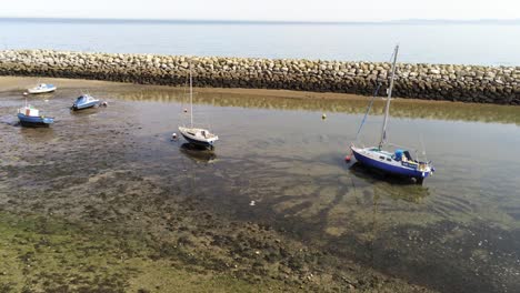 Aerial-view-boats-in-shimmering-low-tide-sunny-warm-Rhos-on-Sea-seaside-rocky-sand-beach-jetty-pull-away-orbit-left