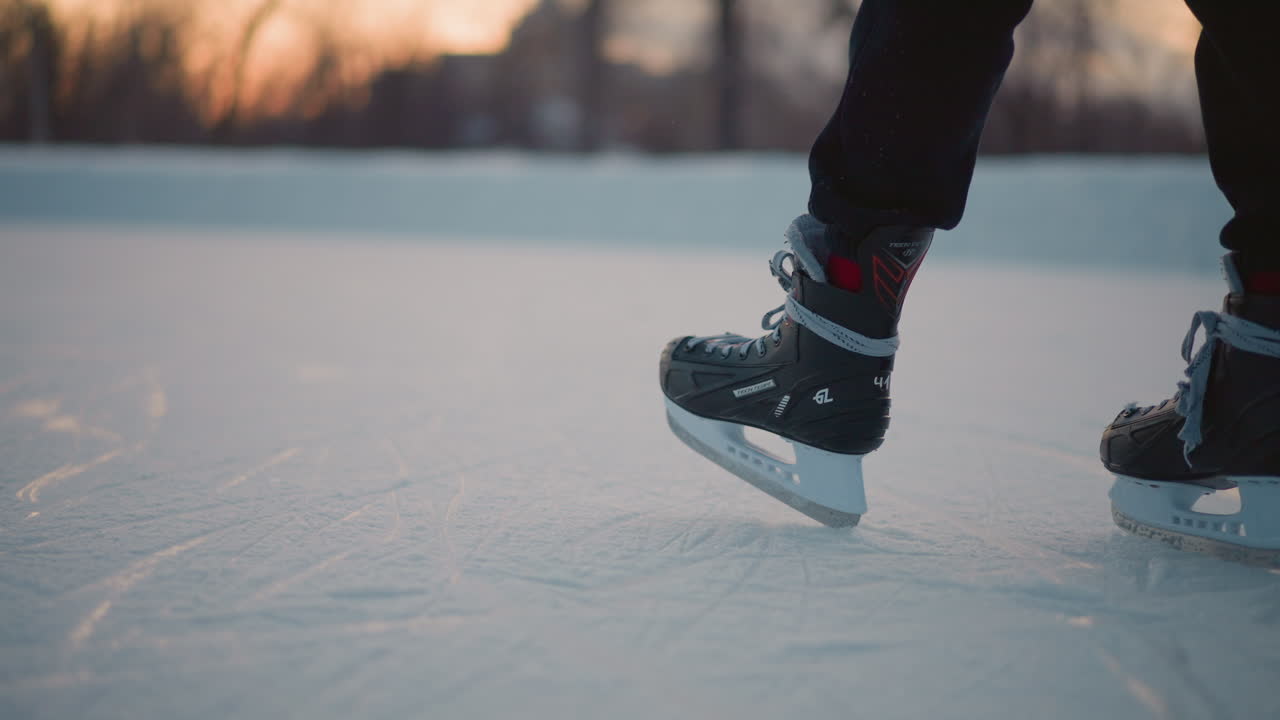 close up leg view skater standing on ice rink in black skating boots laced with gray strings textured icy surface beneath feet with soft sunset light and blurred distant trees in winter background