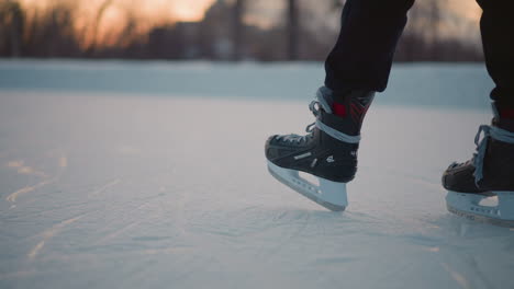 close up leg view skater standing on ice rink in black skating boots laced with gray strings textured icy surface beneath feet with soft sunset light and blurred distant trees in winter background