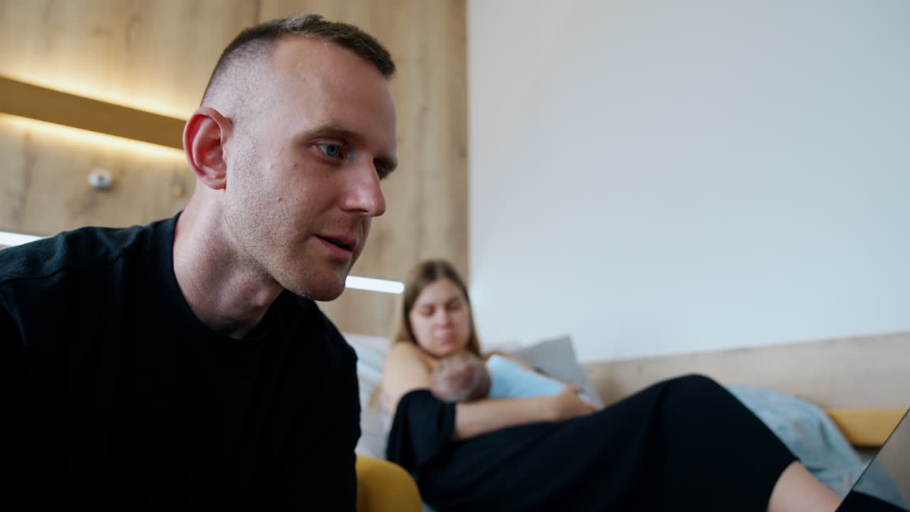 Long-haired woman sitting on the bed is focused on breastfeeding a newborn. Man sits beside in the armchair focused on laptop.
