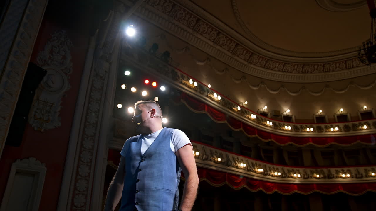 Showman entertains the audience under the light, front view. Big stage. Serious performer speaks from the stage. Hands in pockets.