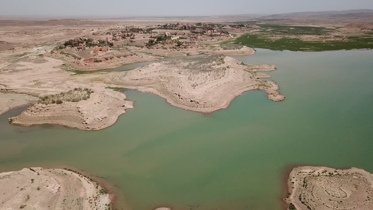 Aerial wide shot of a dam reservoir in Ouarzazate. The turquoise water contrasts with the dry sandy shores, highlighting water storage infrastructure in the Moroccan desert