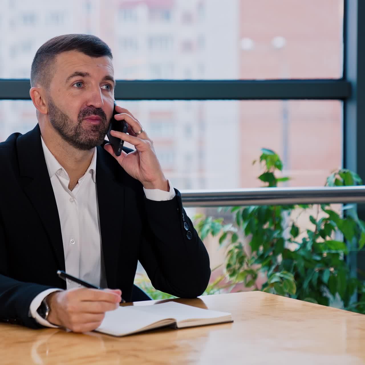 Riunione di lavoro al telefono. Uomo barbuto di mezza età in giacca e cravatta che parla al telefono con dei partner commerciali. Finestra con paesaggio urbano sfocato sullo sfondo