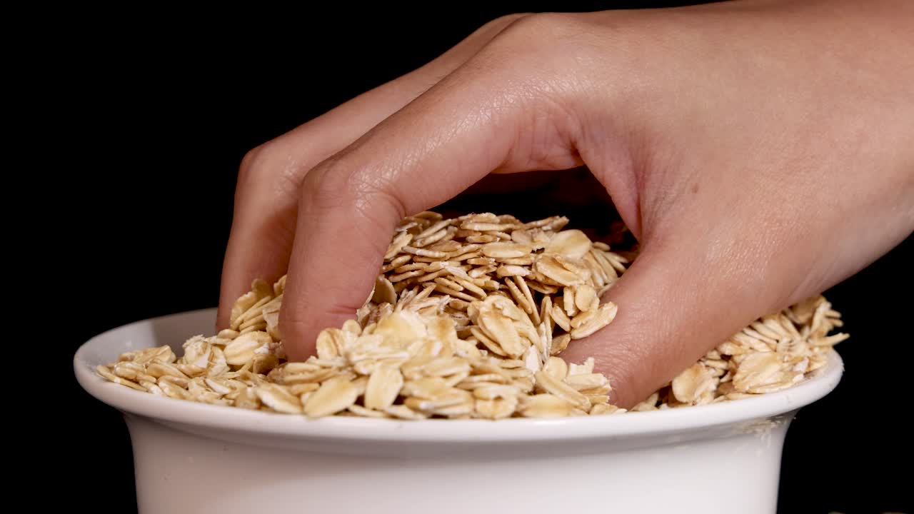 A hand sifts through oats in a bowl against a black background, showcasing tactile interaction and texture