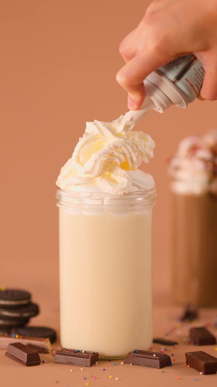 Close-up of hand topping vanilla milkshake with whipped cream, cookies, chocolate, and warm lighting