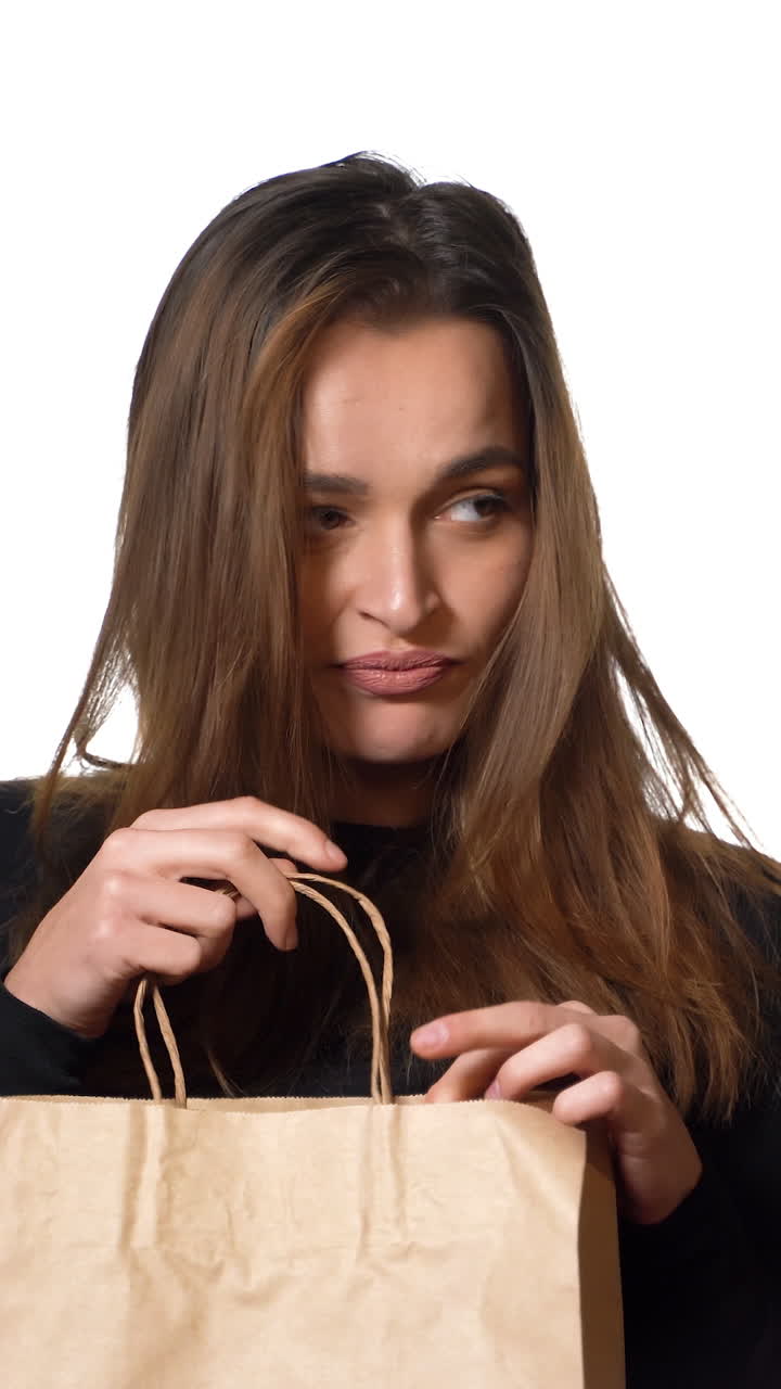 A beautiful woman in a black sweater looks mysteriously in a paper packbag and throws it in frustration and go away on the white background. Purchases. Shopping Vertical video