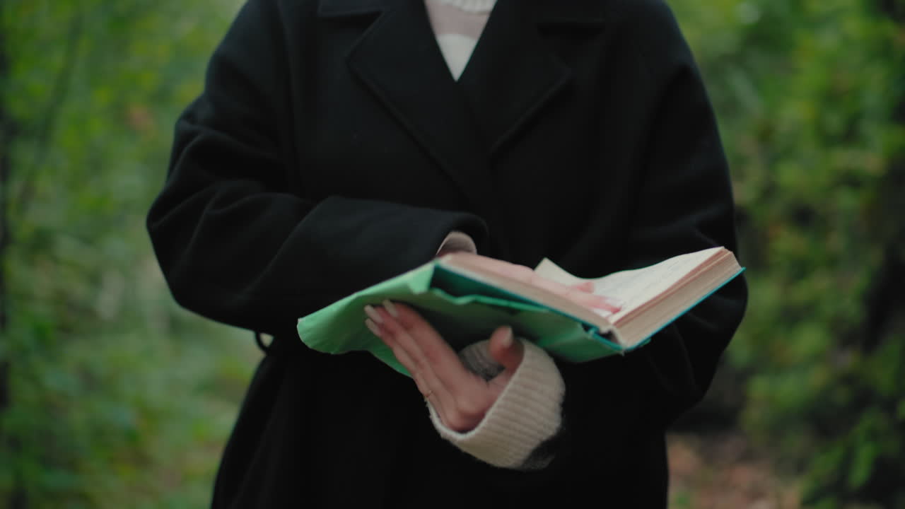 가을 코트를 입은 평화로운 소녀가 숲길을 거닐며 책 페이지를 넘기는 모습, 부드럽게 초점이 맞춰진 주변 녹색 풍경, 야외에서 즐기는 평온한 독서 시간, 아늑한 커프스, 펼쳐진 책과 함께 사색에 잠긴 산책
