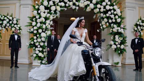 bride making dramatic motorcycle entrance at wedding celebration, wearing elegant white dress and veil, surrounded by supportive groomsmen and vibrant floral decorations