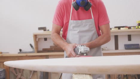 fabricante de tablas de surf masculino caucásico con una máscara facial y preparándose para pulir una tabla de surf