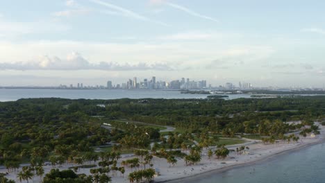 tomada aérea de la hermosa playa tropical rodeada de palmeras en el parque crandon en key biscayne con el horizonte de miami, florida en la distancia en una cálida noche soleada de verano