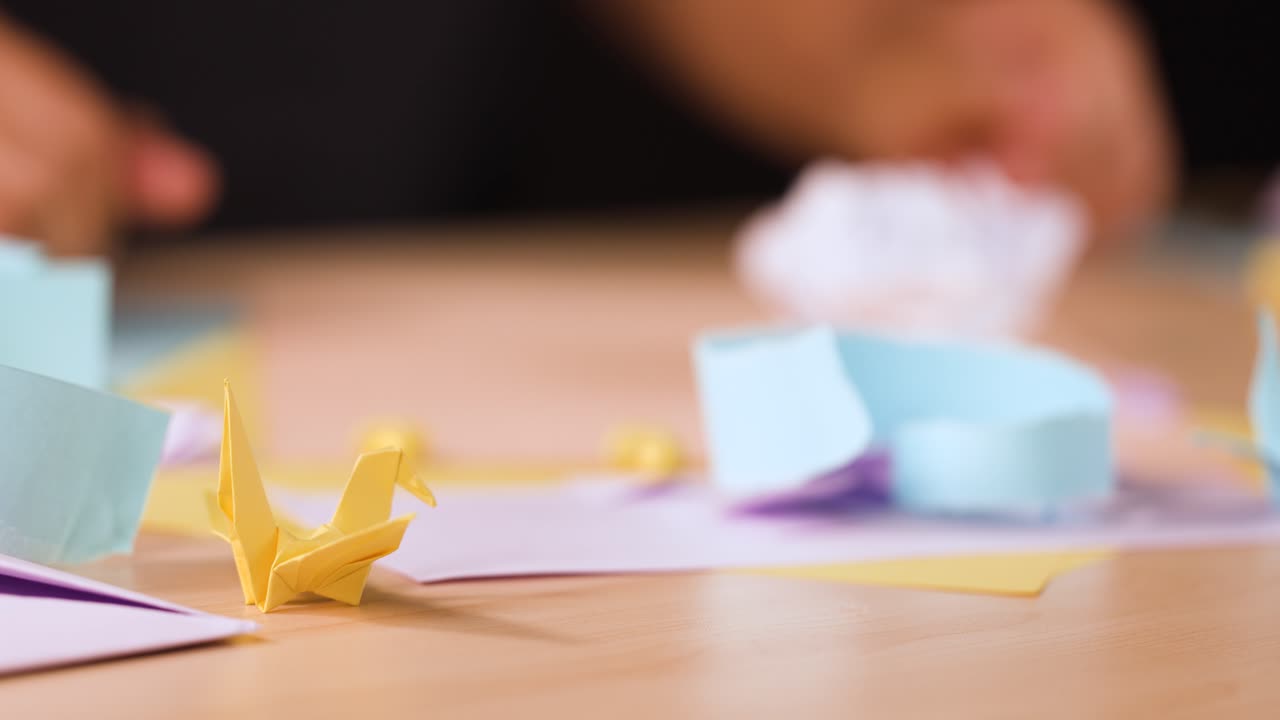 Una persona piega un foglio di carta bianco in forme origami su una scrivania con lavoretti di carta colorati