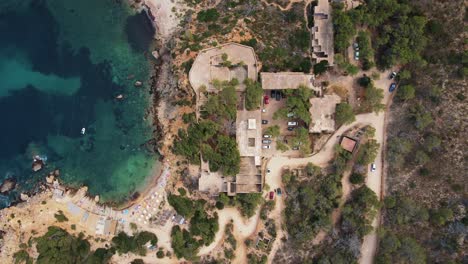 Vista-Aérea-Superior-Del-Hotel-Abandonado-Sert-En-La-Playa-De-Cala-D&#39;en-Serra,-España-Y-Océano-Azul-Claro-Con-Acantilados-Rocosos,-Caminos-Y-árboles