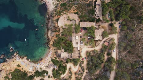 aerial top view of abandoned hotel sert at cala d’en serra beach, spain and clear blue ocean with rock cliffs, roads and trees