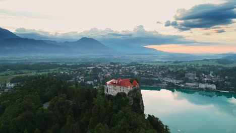 bled castle on rock overlooking beautiful lake bled slovenia and town with mountains in background, aerial parallax drone shot, peaceful travel destination, early morning sunrise, cinematic 4k