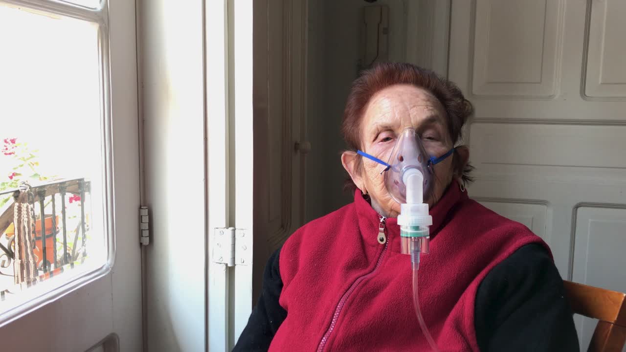 rodeando a una anciana caucásica sentada junto a una ventana con máscara que recibe tratamiento con nebulizador