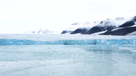 Spectacular-aerial-view-of-a-glacier-with-snow-capped-mountains-in-the-background-in-a-cloudy-day-during-a-boat-expedition-in-the-Arctic-Sea-along-the-northern-coastline-of-Svalbard