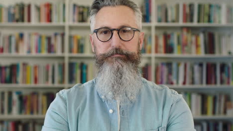 close up portrait of mature caucasian professor with beard looking serious at camera in library wearing glasses bookshelf background