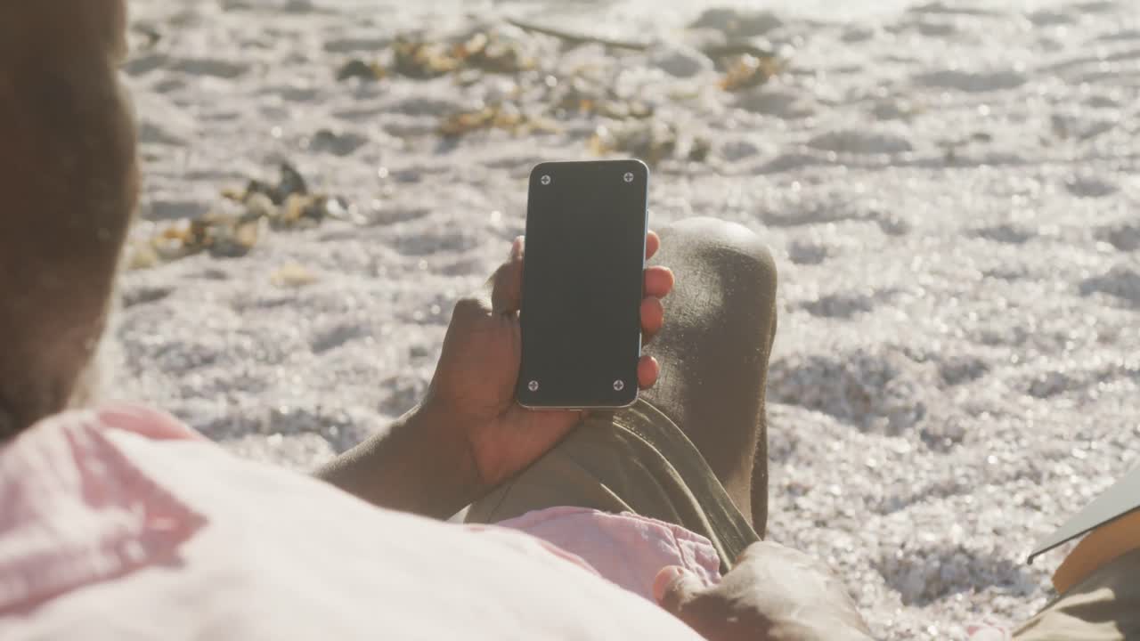 시니어 아프리카계 미국인 남자가  ⁇ 자리에 누워서  ⁇ 은 해변에서 스마트폰을 사용하고 있습니다.