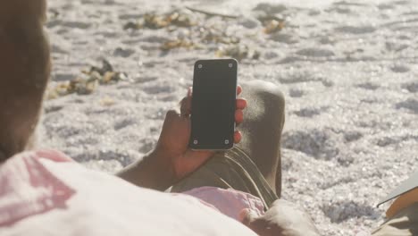 hombre afroamericano de edad avanzada acostado en una cama de sol y usando un teléfono inteligente en una playa soleada