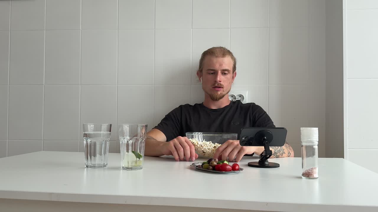 Junger Mann isst Popcorn und Snacks am Küchentisch.
