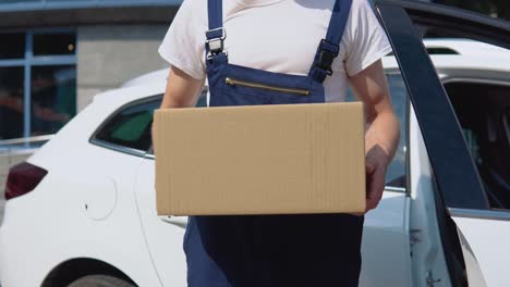 a delivery worker in a white t-shirt and blue jumpsuit stands by an open car and gives a box to recipient in the direction to the camera