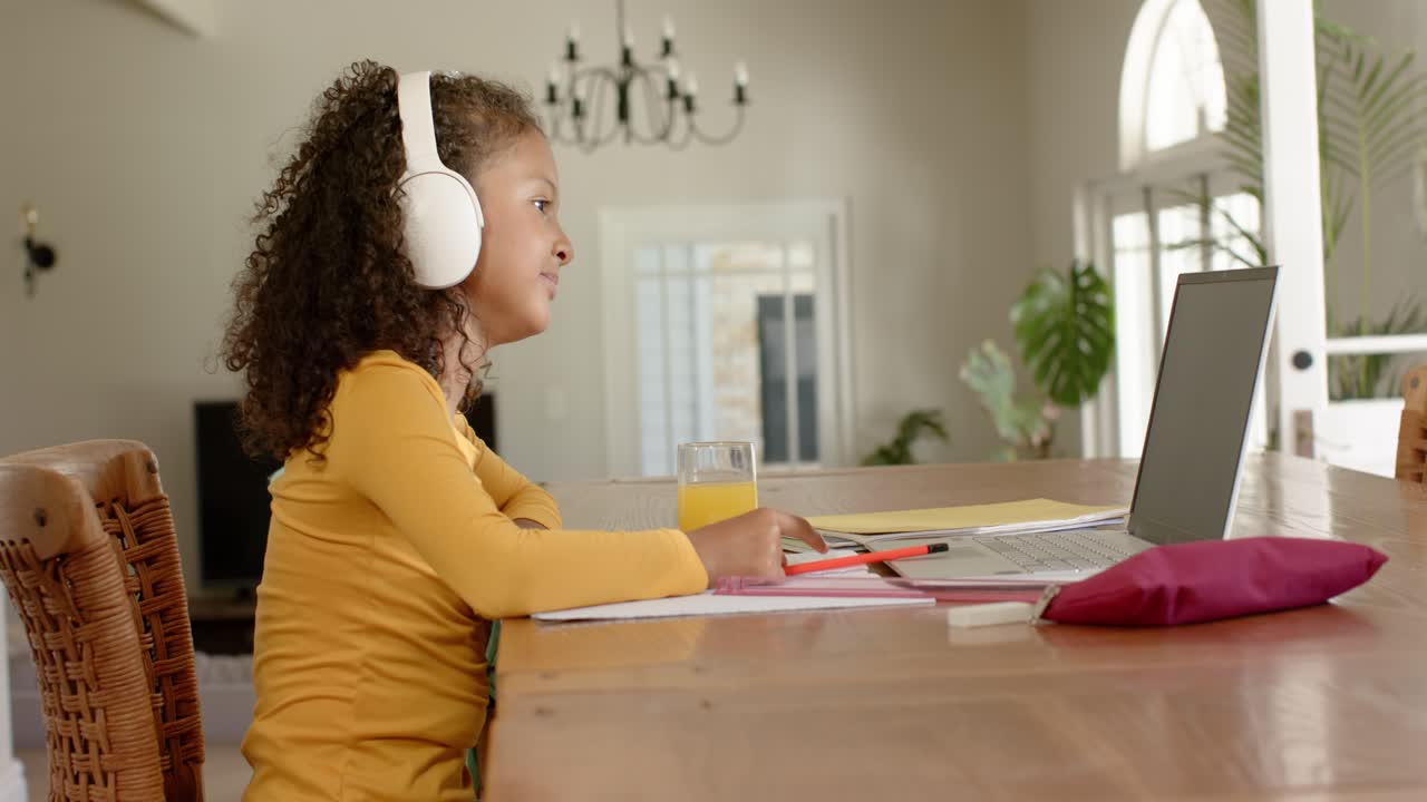 Schoolgirl in yellow shirt writing at laptop on table using headphones sipping juice, copy space