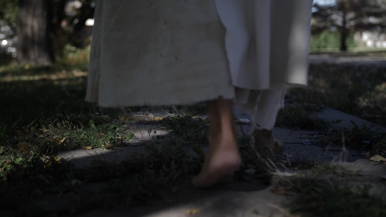 A dramatic slow motion closeup of Jesus' feet walking down a sidewalk while he wears a white, tattered robe