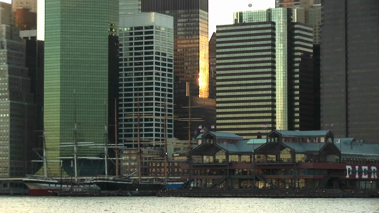 카메라는 골든아워에 해안 쪽 사우스 스트리트 항구에서 엠파이어 스테이트 빌딩을 포함하여 주변 고층 건물의 맨 꼭대기까지 패닝합니다.